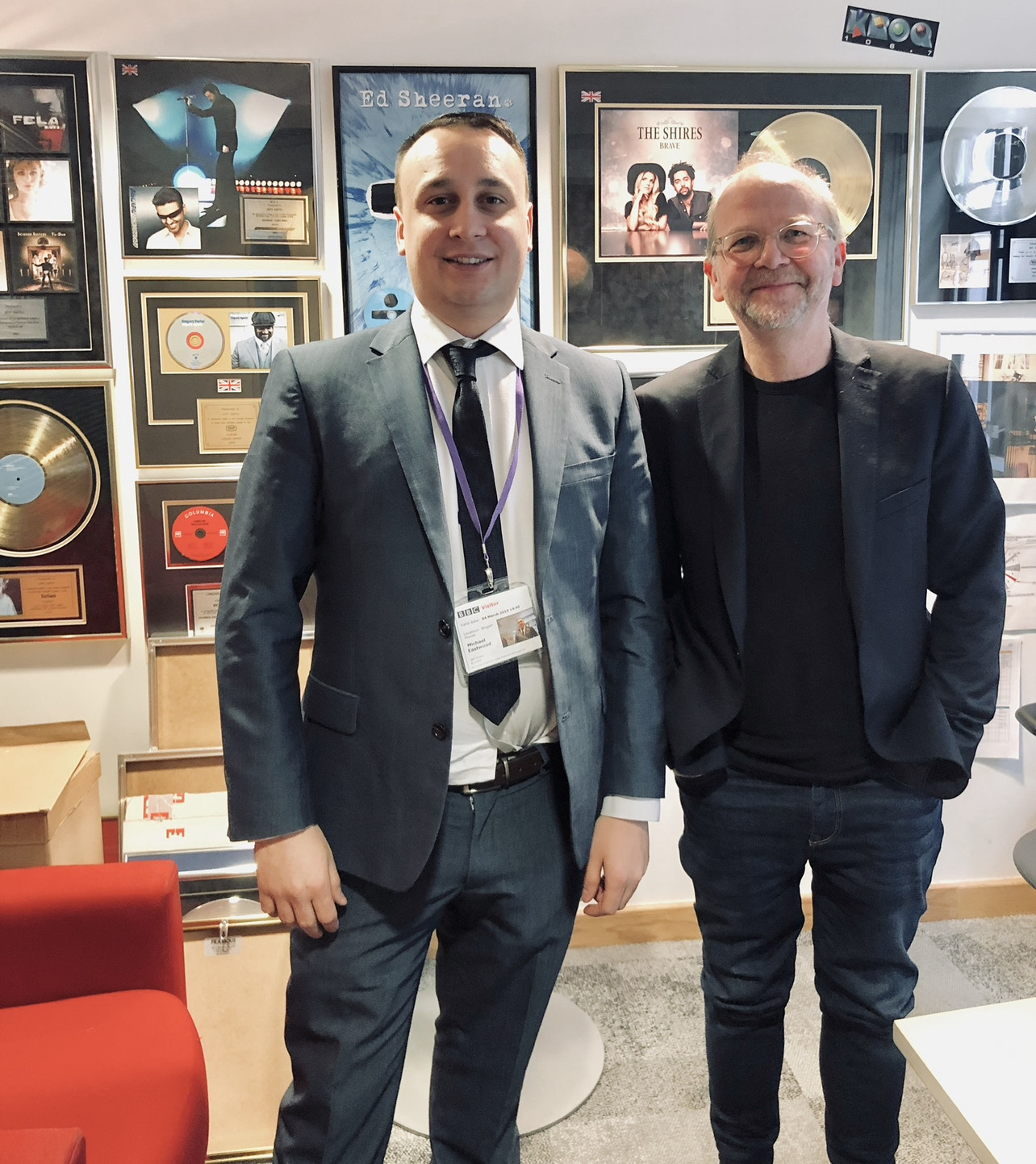 Michael Darius Eastwood wearing a dated BBC visitor pass, standing beside the head of BBC Radio 2 and 6 Music in his office, walls covered with framed gold and platinum discs
