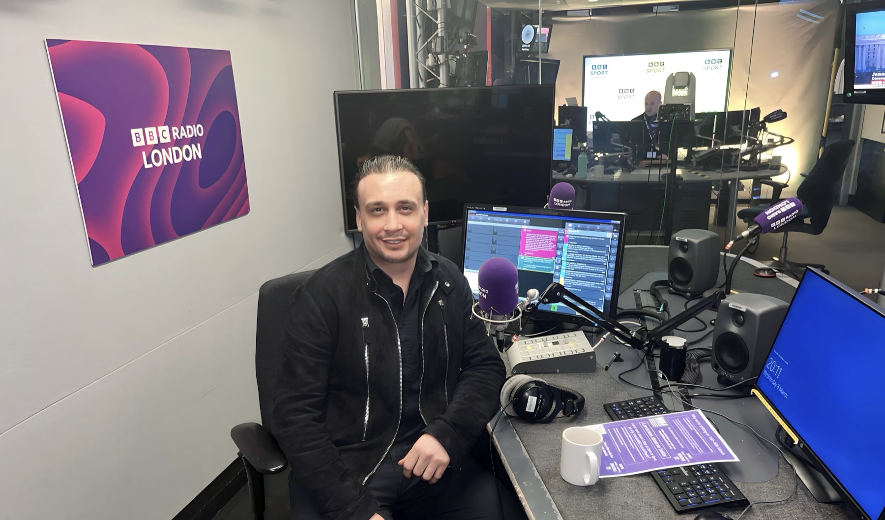 Michael Darius Eastwood seated at the BBC Radio London on-air desk beside a branded microphone, the studio playout screens live and the adjoining BBC Sport studio visible through the glass
