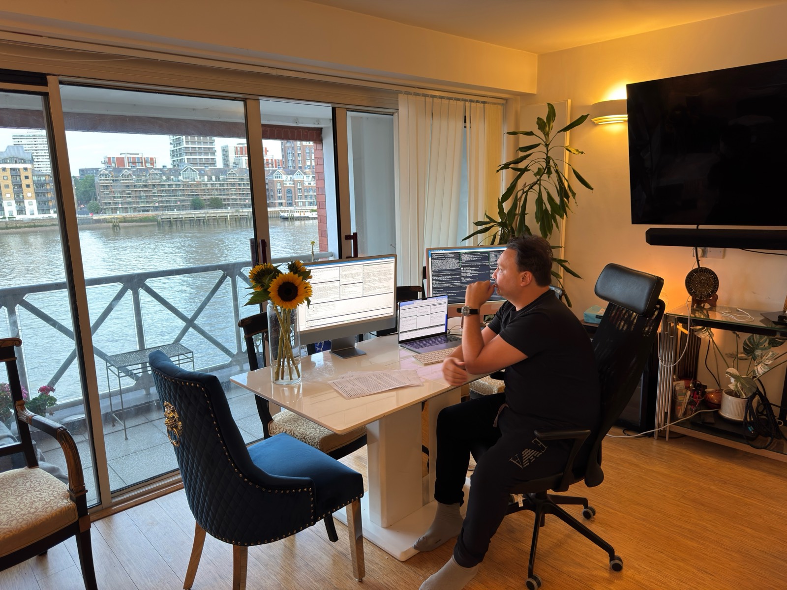 Michael Darius Eastwood working at a table by a window over the Thames, three screens running, a laptop between them, papers and sunflowers on the table