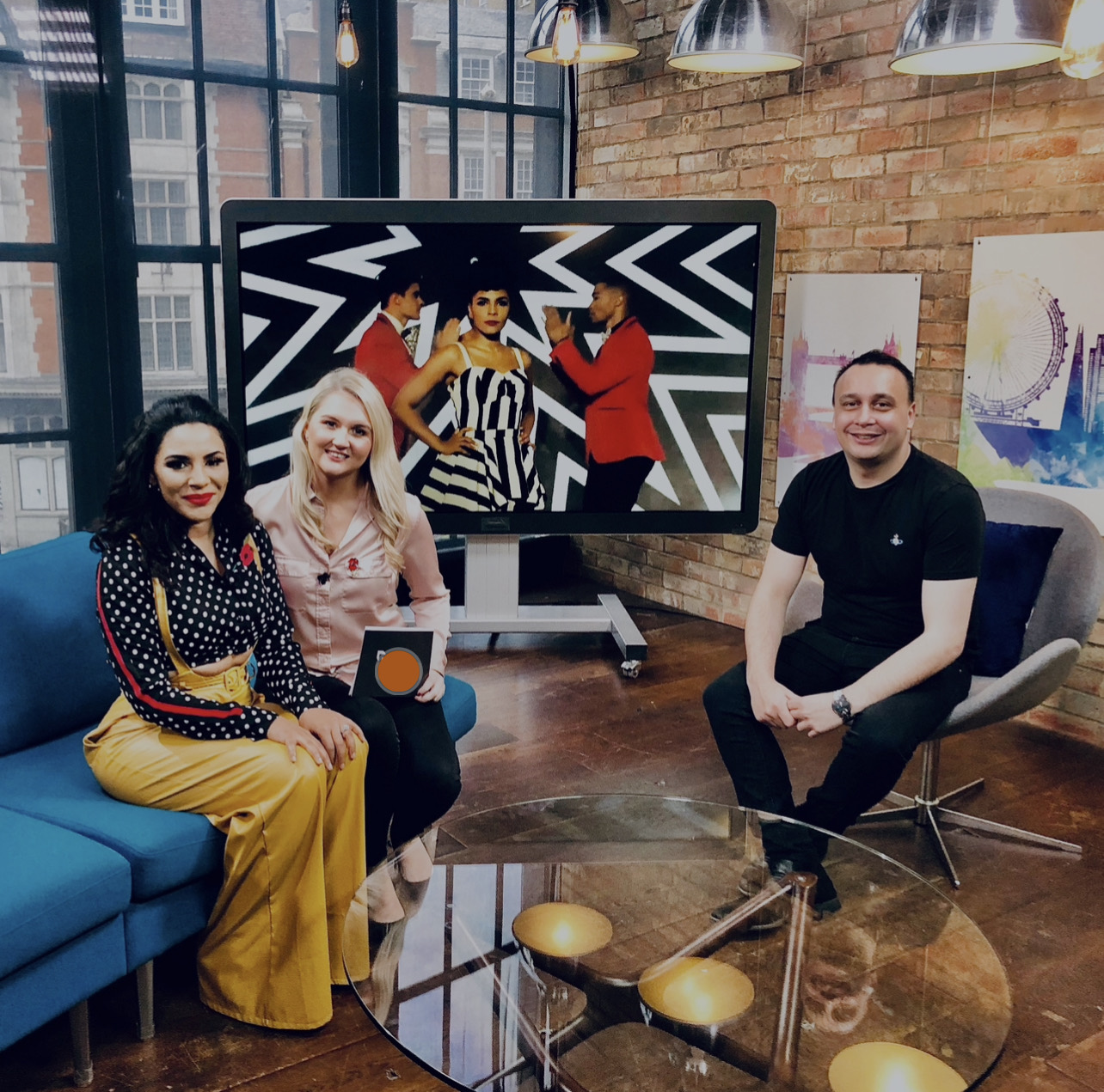 Michael Darius Eastwood seated in the presenter chair on the London Live television set before broadcast, with two presenters on the sofa and a music video on the studio screen