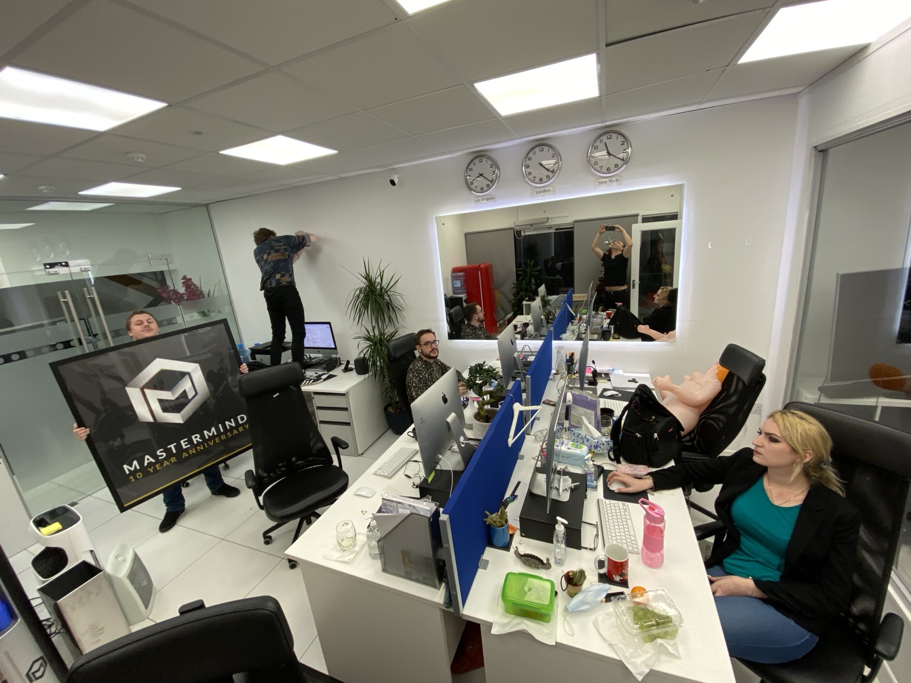 The company office at work, a staff member carrying a large framed Mastermind ten year anniversary board, another standing on a desk to hang it, colleagues working at iMacs, three clocks labelled Los Angeles, London and New York