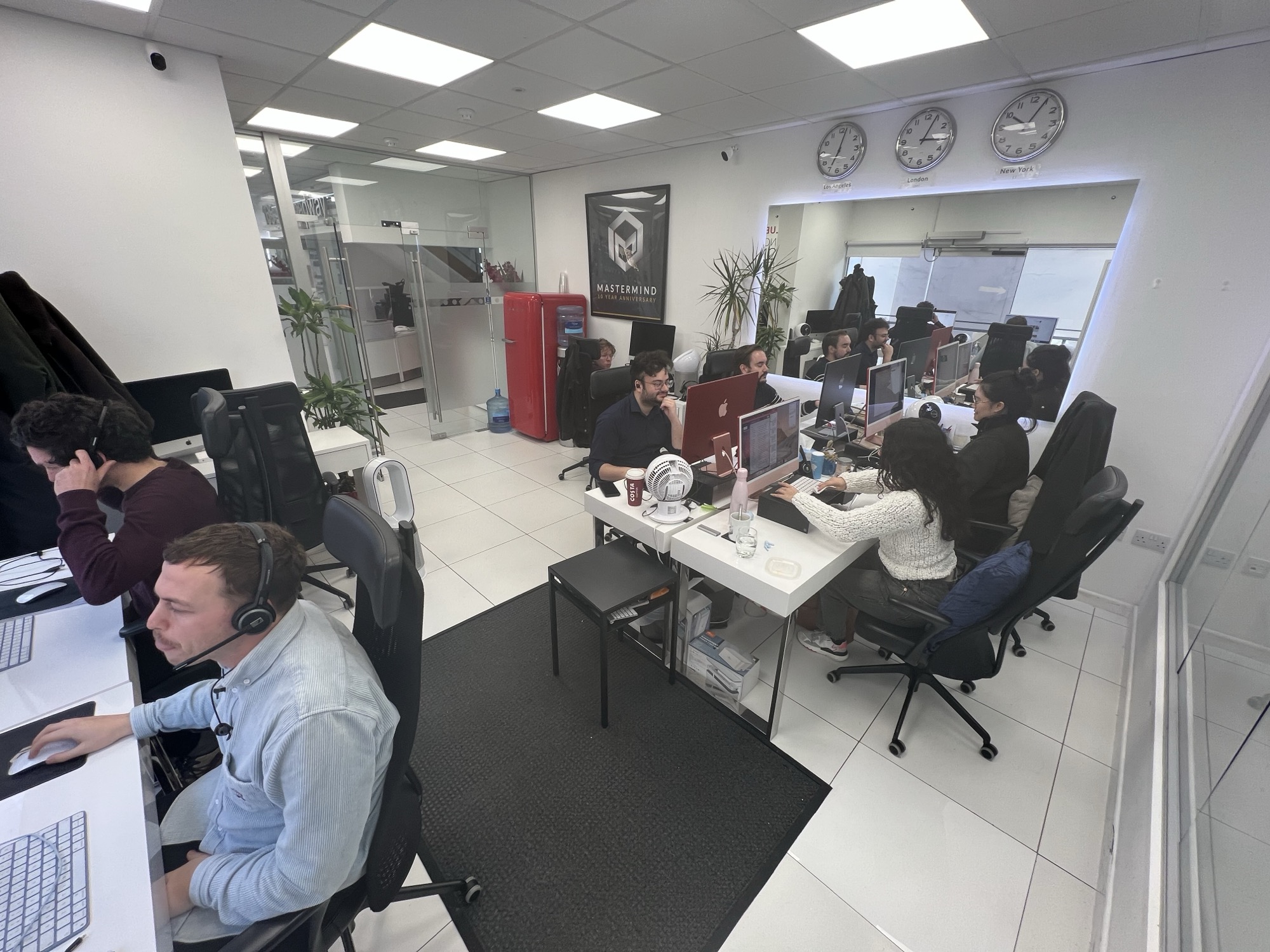 The company floor in London: about ten people at iMacs, several wearing telephone headsets, three wall clocks labelled Los Angeles, London and New York, and a framed ten-year anniversary poster