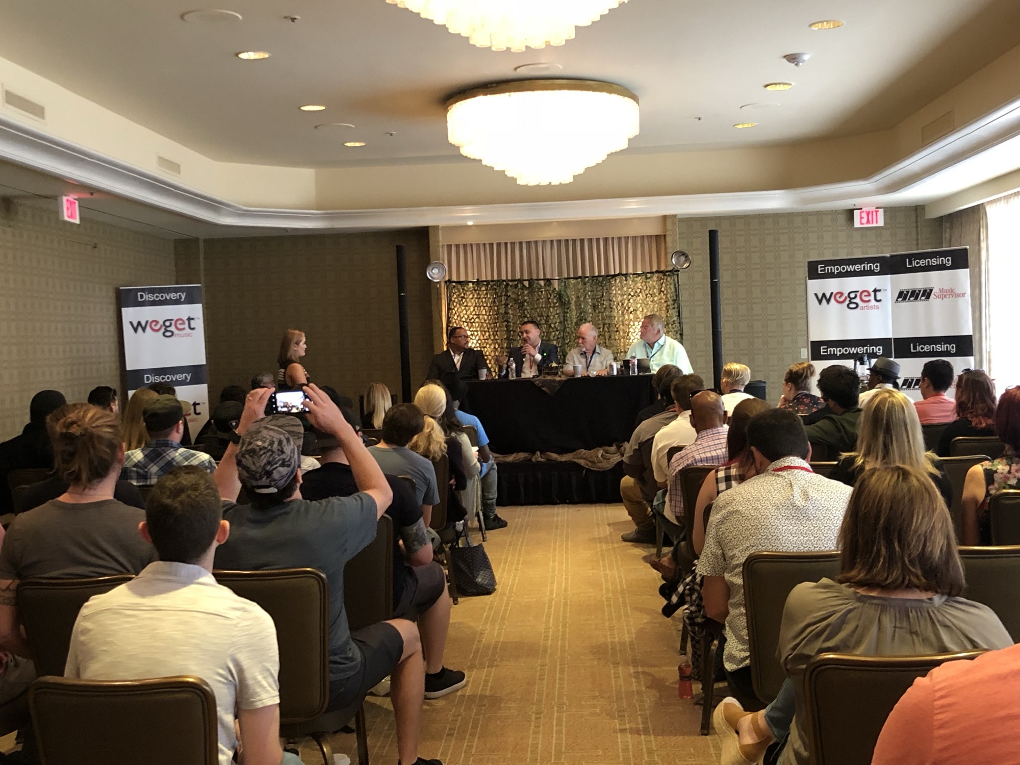 Michael Darius Eastwood speaking with a microphone on a four-person industry panel at a music conference, a full room of delegates seated in front of the stage